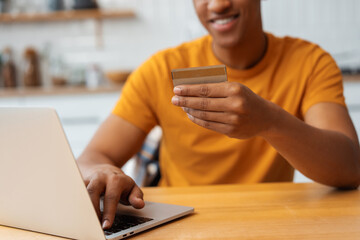 Smiling authentic African American young man using laptop, holding credit card, ordering, buying