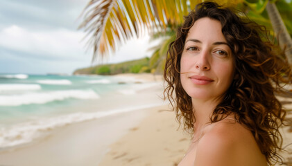 Beautiful mature woman sitting on a sandy beach under palm trees against the background of the sea, woman looking at the camera, curly hair fluttering in the wind, resort holiday concept