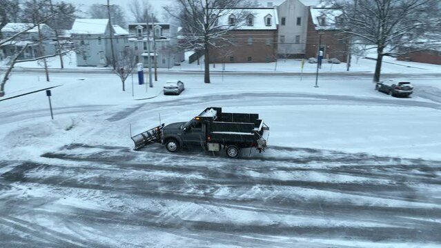 Pickup truck with dump bed and snow plow clearing snow covered parking lot. Aerial tracking shot during snow storm in winter in USA.