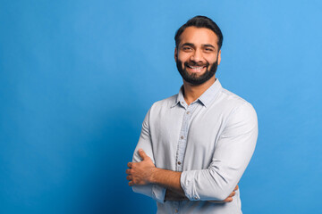 Fototapeta premium In front of a bright blue background, the Indian businessman stands with arms crossed, his casual light blue shirt suggesting a relaxed yet confident business approach
