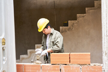 Young male bricklayer taking mortar with trowel from bucket while building brick wall at incomplete...