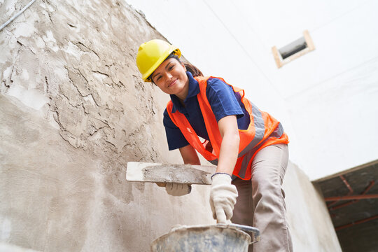 Low angle portrait of smiling young female bricklayer plastering on wall with cement at construction site