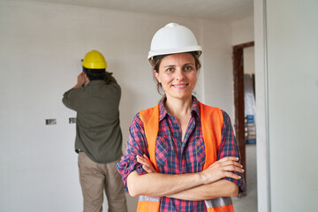 Portrait of smiling young female builder standing with arms crossed against coworker at incomplete...