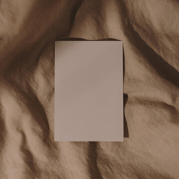 Blank paper card sheet with copy space on wrinkled bed blanket cloth with aesthetic warm sunlight shadows. Flat lay, top view