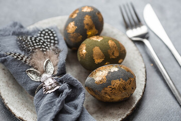 able setting. A fashionable concrete plate with a rabbit on a napkin, Easter eggs with gilding, feathers on a gray background. The concept of a Happy Easter holiday for cafes and restaurants.