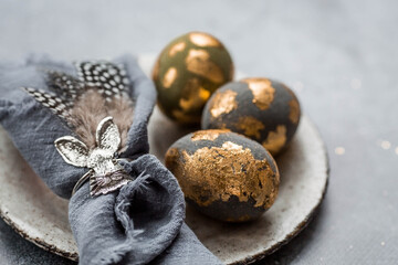 able setting. A fashionable concrete plate with a rabbit on a napkin, Easter eggs with gilding, feathers on a gray background. The concept of a Happy Easter holiday for cafes and restaurants.