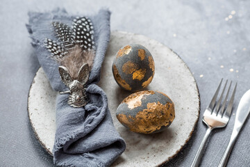 able setting. A fashionable concrete plate with a rabbit on a napkin, Easter eggs with gilding, feathers on a gray background. The concept of a Happy Easter holiday for cafes and restaurants.
