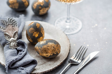 able setting. A fashionable concrete plate with a rabbit on a napkin, Easter eggs with gilding, feathers on a gray background. The concept of a Happy Easter holiday for cafes and restaurants.