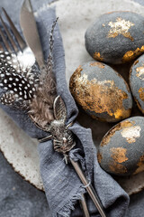 able setting. A fashionable concrete plate with a rabbit on a napkin, Easter eggs with gilding, feathers on a gray background. The concept of a Happy Easter holiday for cafes and restaurants.