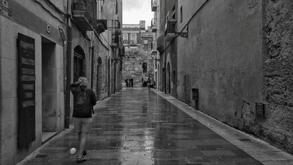a woman walking down a narrow street in a city - obrazy, fototapety, plakaty