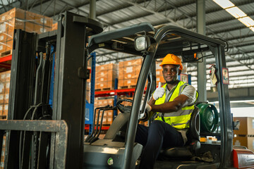 African forklift driver focused on carefully transporting stock from shelves of a large warehouse wearing a helmet and vest looking toward goods