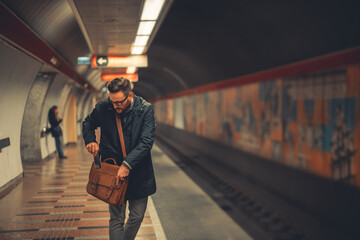 An adult businessman putting away his phone in his bag over his shoulder as he waits for the train - obrazy, fototapety, plakaty