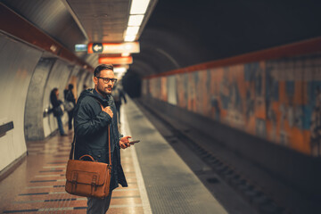 A portrait of a serious businessman with a shoulder bag waiting for the train on a subway station - obrazy, fototapety, plakaty