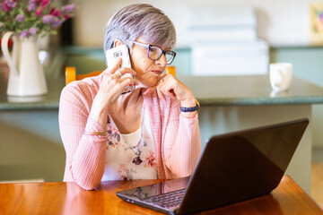 Mature woman on laptop at home using mobile phone to call for tech support