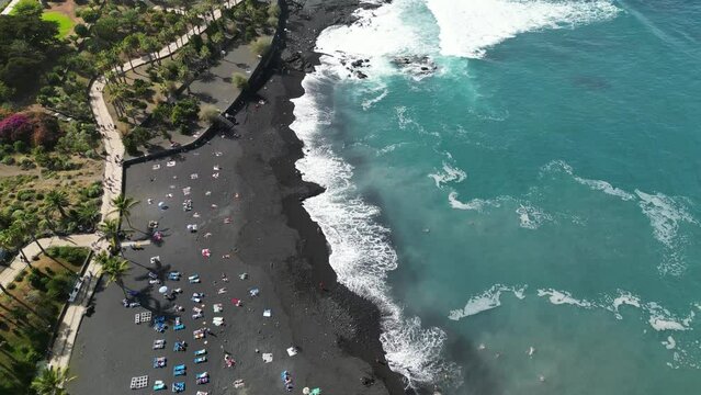 Aerial view of central beach in Puerto de la Cruz on the Canary Islands. Drone flying above Jardin beach. Beautiful black volcanic sand beach with palms and blue water.