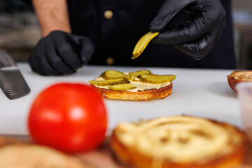 The cook in the kitchen is cooking a cheeseburger and putting a pickled cucumber.
