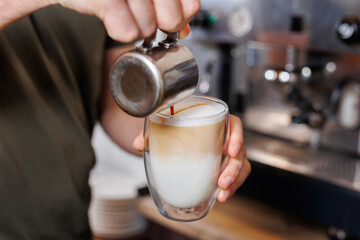 Close-up of barista gently drawing pattern pouring milk into glass with latte.