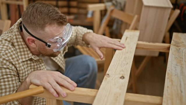 Focused man with beard examines chair in workshop wearing safety goggles