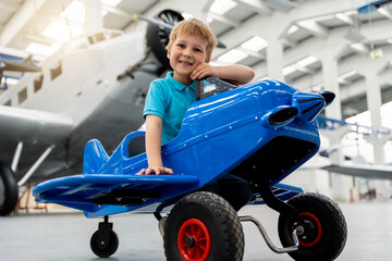 Portrait of cute little blond happy kid boy enjoy have fun play riding pedal toy plane model against vintage airplane background in museum hangar. Future child profession dream concept