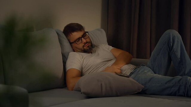 Tired man in glasses and clothes sleeps on sofa with phone in evening at home after hard day. Man naps with smartphone