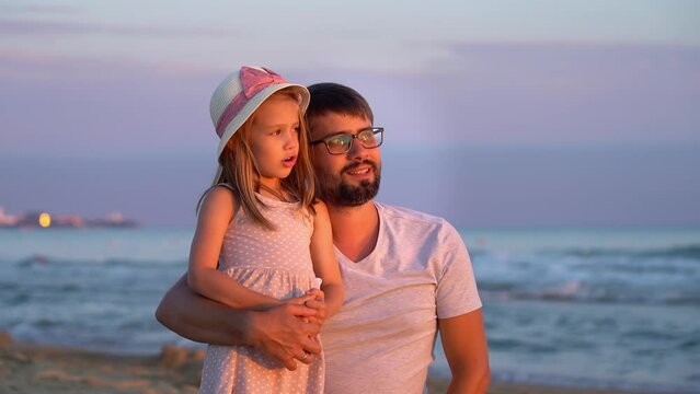 Happy family, father and daughter sit on beach with blue sea and look at sunset. Concept of summer vacation, travel, holiday. People outdoors on seascape