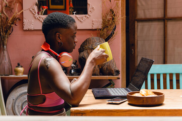 Side view of young afro woman drinking coffee and working with laptop, calm at home.