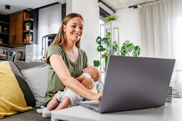 Young new mom holding cute baby in her arms at home while working on a computer - obrazy, fototapety, plakaty