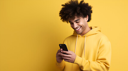 A young smiling curly man in the yellow hoodie holding a phone on the yellow background
