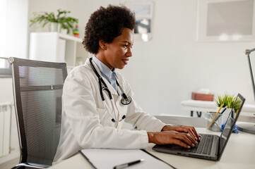 Female doctor sitting at her office in front of laptop computer doing consultations online over internet.