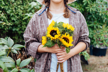 Focus on fresh sunflowers bouquet in hands of young female farmer on the green trees background....