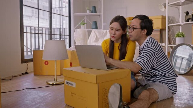 Asian Cheerful young married couple sitting on floor during enjoy unpack home relocation moving in new home or apartment,having coffee break and arrange decorative items on shelf.home moving concept