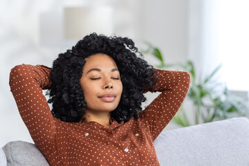 Tired young African American woman resting at home, sitting relaxed on sofa with hands behind head...