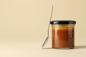 Tasty salted caramel in jar and spoon on pale yellow background, space for text