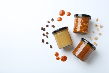 Tasty salted caramel in jars with peanuts and coffee beans on white background, flat lay. Space for...