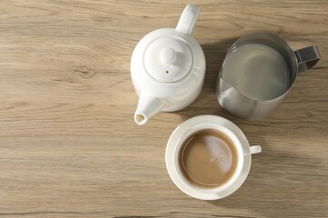 Aromatic tea with milk in cup, teapot and pitcher on wooden table, flat lay. Space for text