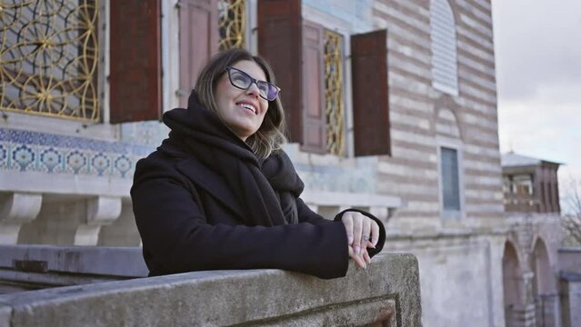 Smiling woman wearing glasses and a black coat leans on a historic balcony in istanbul, with traditional ottoman architecture in the background.