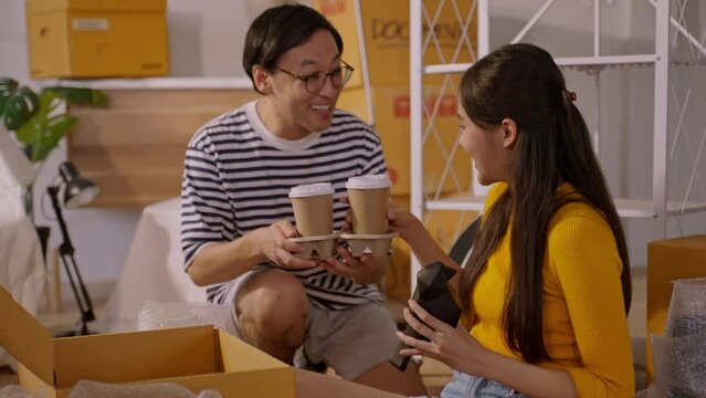 Asian Cheerful young married couple sitting on floor during enjoy unpack home relocation moving in new home or apartment,having coffee break and arrange decorative items on shelf.home moving concept