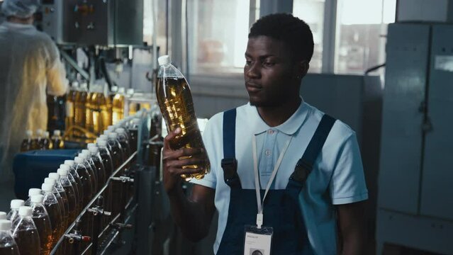 Medium stab footage of Black manufacturing engineer controlling quality of soda production line standing with plastic bottle near conveyor belt at beverage plant