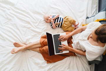 Mother working at laptop playing with baby in bed at home.