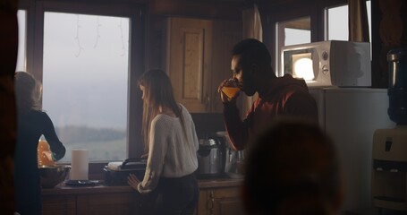 Multiethnic group of friends cook breakfast together in holiday house. African American man stands in kitchen, drinks juice. Big travelers family stopped to rest in cottage during vacation. Tourism.