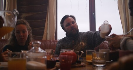 Big multicultural family sit at table, eat, drink, talk. Adult man pours milk to plates. Group of...