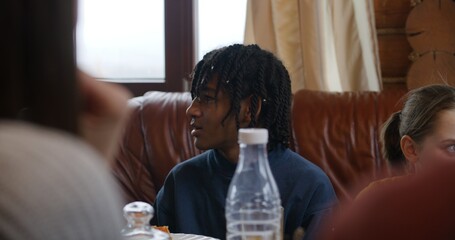 Close up shot of African American teenager pointing finger up. Big multi ethnic family sit at table, eat, drink and talk. Group of friends gathered in holiday house. Family leisure together concept.