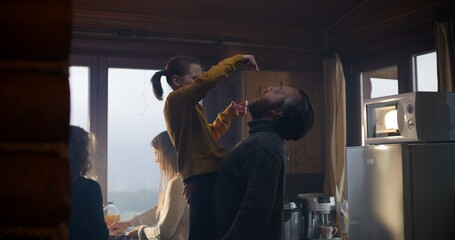 Adult man stands in kitchen, takes on hands daughter and eats with her. Women cook breakfast together in holiday house. Big travelers family stopped to rest in cottage during vacation. Family leisure.