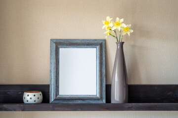 Vintage interior decor with a bouquet of yellow daffodils in a vase, candle holder and an antique empty blue frame on a vintage shelf over textured beige wall. Home decor on a shelf.