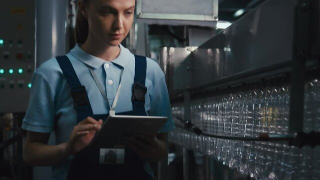 Tilt footage of young Caucasian female inspector examining plastic bottles manufacturing process using tablet and walking along conveyor line at mineral water plant