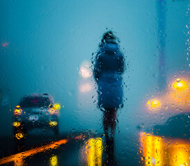 View through a glass window with raindrops on a blurred silhouette of a girl on a autumn city street after rain against the bokeh of  city lights, night street scene. Focus on raindrops on glass	