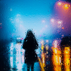 View through a glass window with raindrops on a blurred silhouette of a girl on a autumn city street after rain against the bokeh of  city lights, night street scene. Focus on raindrops on glass	