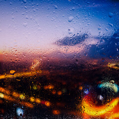  View through a glass window with raindrops on city streets with cars in the rain, bokeh of colorful city lights, night street scene. Focus on raindrops on 