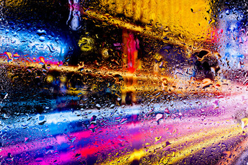 View through a glass window with raindrops on city streets with cars in the rain, bokeh of colorful city lights, night street scene. Focus on raindrops on glass