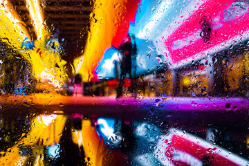  View through a glass window with raindrops on city streets with cars in the rain, bokeh of colorful city lights, night street scene. Focus on raindrops on 
